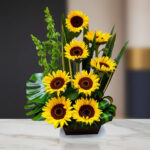 Arreglo floral moderno con varios girasoles amarillos y follaje verde en base rectangular negra sobre mesa de mármol