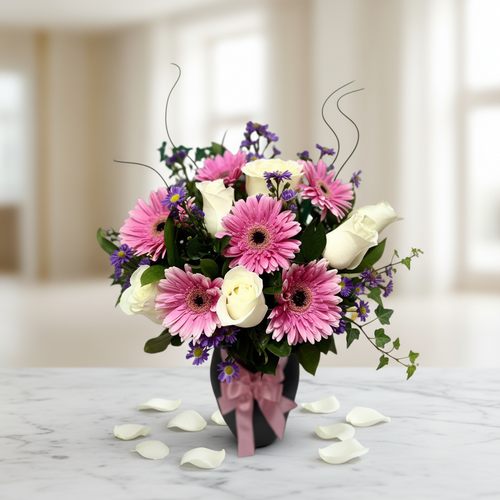 Arreglo floral con gerberas rosas, rosas blancas y pequeñas flores moradas en jarrón negro con lazo rosa sobre mesa de mármol, con pétalos blancos alrededor y fondo interior desenfocado