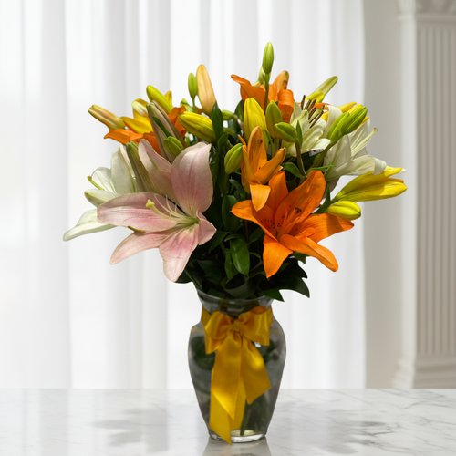 Arreglo de lirios naranjas, amarillos, blancos y rosados en un jarrón de vidrio con lazo amarillo sobre una mesa de mármol