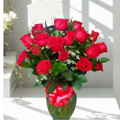 Arreglo de rosas rojas frescas con follaje verde y pequeñas flores blancas en un jarrón de cristal con lazo rojo y tarjeta, sobre una mesa blanca junto a una ventana