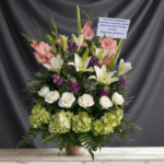 Arreglo floral grande con rosas blancas, lirios blancos, hortensias verdes, flores rosadas y moradas, con tarjeta de condolencias sobre fondo gris