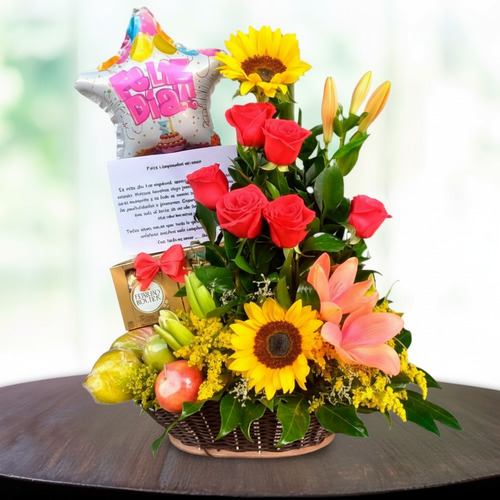 Canasta de mimbre con arreglo floral de rosas rojas, girasoles y lirios naranjas, acompañada de frutas, globo de cumpleaños en forma de estrella y tarjeta sobre una mesa redonda