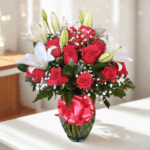 Arreglo floral con rosas rojas, lirios blancos y paniculata en jarrón de cristal con lazo rojo sobre mesa blanca