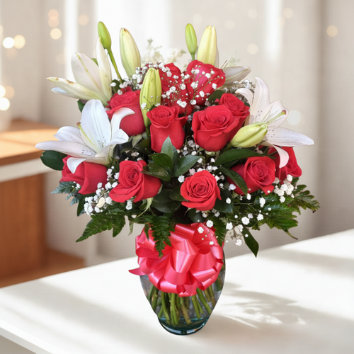 Arreglo floral con rosas rojas, lirios blancos y paniculata en jarrón de cristal con lazo rojo sobre mesa blanca