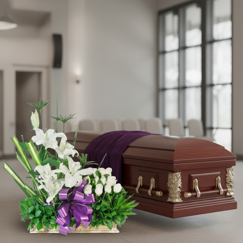 Ataúd de madera marrón con detalles dorados y tela púrpura, junto a un arreglo floral de lirios y rosas blancas con lazo morado en una sala funeraria luminosa