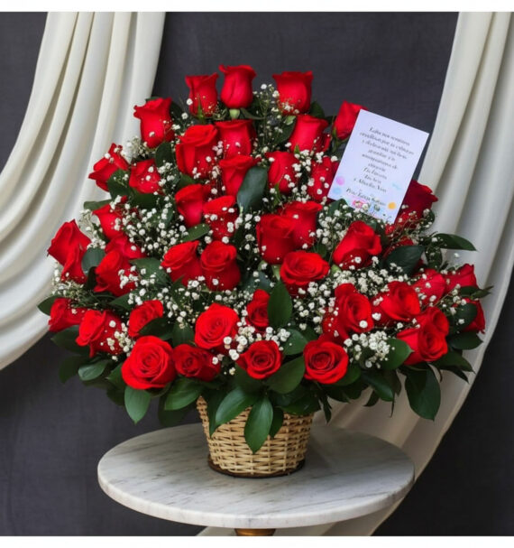 Canasta de mimbre sobre mesa redonda con gran arreglo de rosas rojas y flores blancas de relleno, acompañadas de tarjeta sobre fondo de cortinas blancas y tela gris