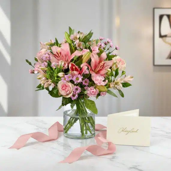 Arreglo floral ANNA con rosas rosadas, lirios y alstroemerias en jarrón de cristal, sobre mármol blanco con tarjeta de regalo