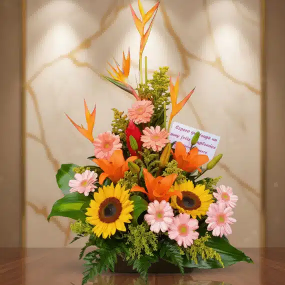 Arreglo Floral Exótico Columba: Belleza Tropical para Celebraciones Especiales Arreglo floral exótico Columba con aves del paraíso, girasoles, lirios naranjas y margaritas rosadas sobre una mesa de madera