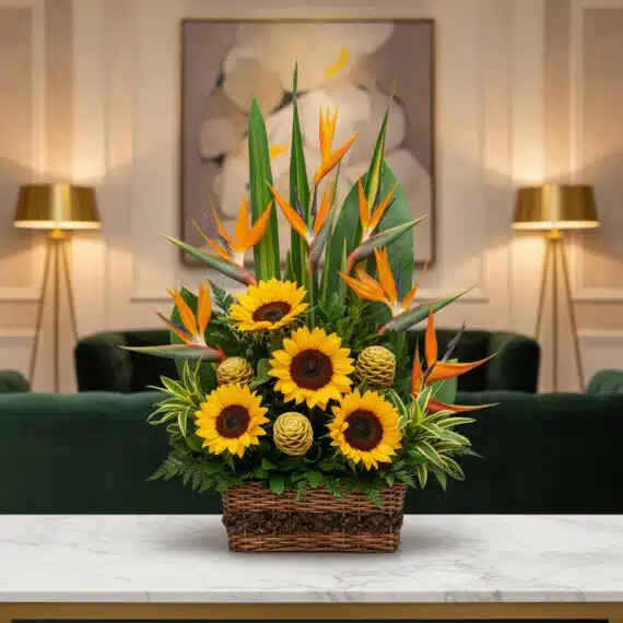 Arreglo Floral Fantasía: Girasoles y Aves del Paraíso para tu Hogar Arreglo floral Fantasía con girasoles vibrantes, exóticas aves del paraíso y detalles verdes en cesta de mimbre, sobre mesa d