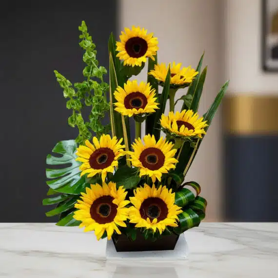 Arreglo Floral con Girasoles Bengala: Elegancia y Frescura para tu Hogar Arreglo floral premium con girasoles Bengala, hojas verdes y follaje, sobre una mesa de mármol en un lobby de hotel boutique.