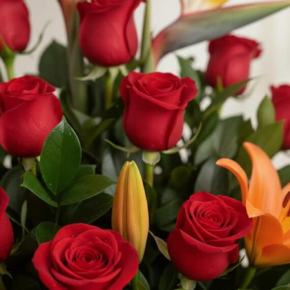 Primer plano extremo de un arreglo floral 'Me Encantas' con rosas rojas, lirios naranjas y follaje verde, gotas de rocío suti