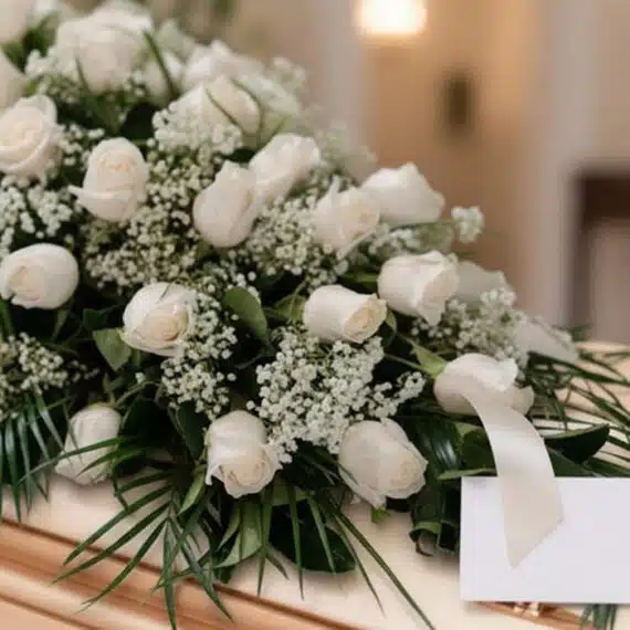 Arreglo fúnebre cubre caja con rosas blancas, follaje verde y pequeñas flores blancas, con una cinta satinada y una tarjeta d