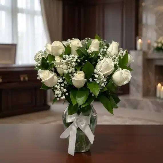 Arreglo Fúnebre Pétalos del Alma: Elegancia y Respeto en Flores Blancas Arreglo fúnebre Pétalos del Alma con rosas blancas y follaje verde en un jarrón de cristal con lazo blanco, sobre una mesa de