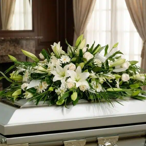 Elegante Arreglo Fúnebre Cubre Caja con Rosas Blancas y Lirios Arreglo fúnebre cubre caja con rosas blancas, lirios y follaje verde, presentado en una sala de velación premium con luz natu