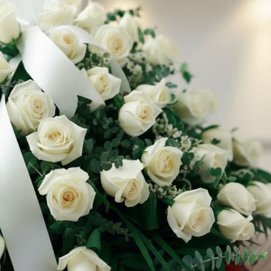 Primer plano de arreglo fúnebre cubre caja con rosas blancas frescas, follaje verde y rocío sutil. Cinta satinada blanca y pr