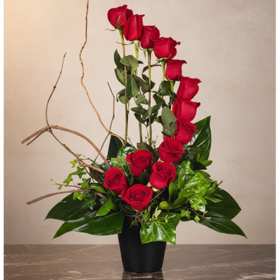 Arreglo floral GIANNA con 13 rosas rojas frescas en un jarrón negro, ramas decorativas y follaje verde, sobre una superficie