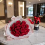Elegante bouquet de 18 rosas rojas abiertas con empaque coreano, caja de chocolates y botella de vino tinto, sobre mesa de má