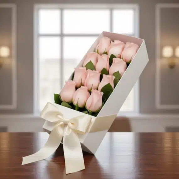 Caja de 12 Rosas Rosadas: El Regalo Perfecto para Cualquier Ocasión Caja blanca inclinada con 12 rosas rosadas frescas y un moño de satén, sobre una mesa de madera oscura con un fondo elegante