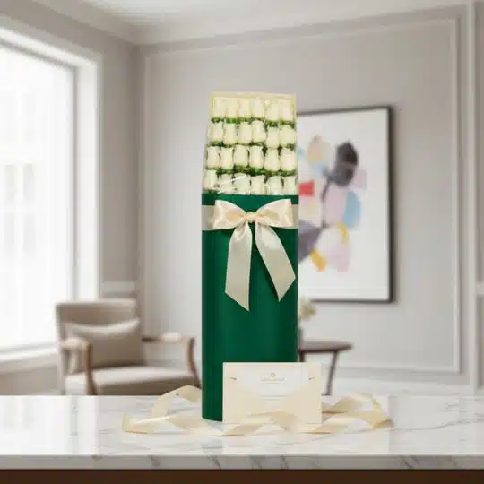 Caja cilíndrica verde con 24 rosas blancas, lazo satinado color crema, tarjeta de regalo y cinta sobre mármol.