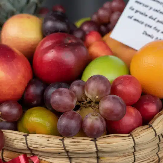 Primer plano extremo de la Canasta de Frutas Musca, mostrando uvas, manzanas, naranjas y otras frutas frescas con gotas de ro