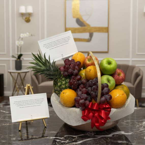 Canasta de frutas premium con piña, uvas, manzanas rojas y verdes, naranjas, y un lazo rojo, sobre una mesa de mármol en un e