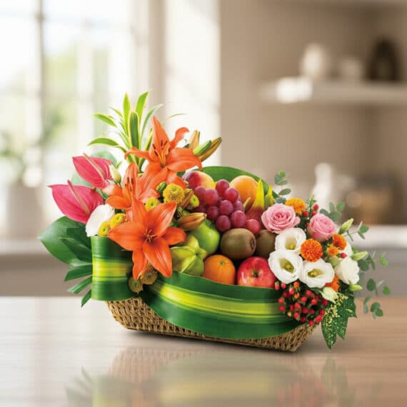 Cesta de frutas THALIA con lirios naranjas, anturios rosas, rosas, uvas, manzanas, kiwis y naranjas, sobre mesa de madera cla