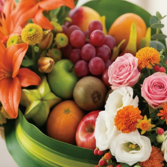 Primer plano extremo de la Cesta Frutal THALIA con lirios naranjas, rosas rosadas, uvas rojas, manzanas, kiwi y carambola, de