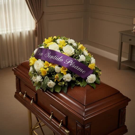 Cubre caja fúnebre Baltasar con rosas blancas, fresias amarillas y follaje verde, adornado con una cinta morada que dice 'Con