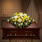 Cubre Caja Fúnebre Baltasar con rosas blancas, freesias amarillas y margaritas, sobre un ataúd de madera oscura en un entorno