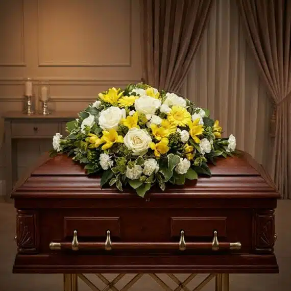 Cubre Caja Fúnebre Baltasar con rosas blancas, freesias amarillas y margaritas, sobre un ataúd de madera oscura en un entorno
