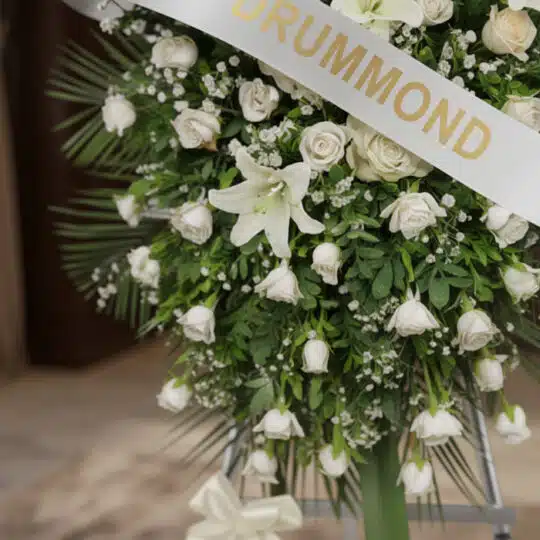 Pedestal conmemorativo Legado de Marcos con arreglo floral fúnebre en tonos blancos y cremas, rosas, lirios y follaje verde,