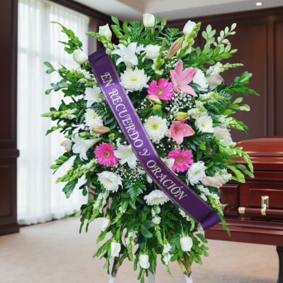 Pedestal fúnebre sagrado con flores blancas, rosas y verdes, con una cinta morada que dice 'En Recuerdo y Oración', junto a u