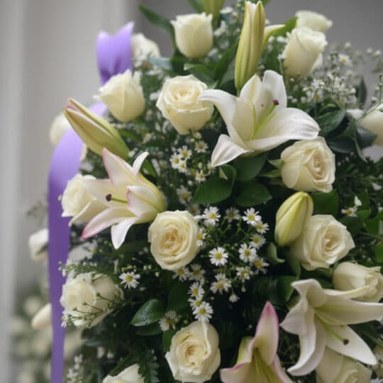 Primer plano de un pedestal fúnebre 'Salvación' con rosas blancas, lirios y follaje verde, destacando texturas y rocío sutil.